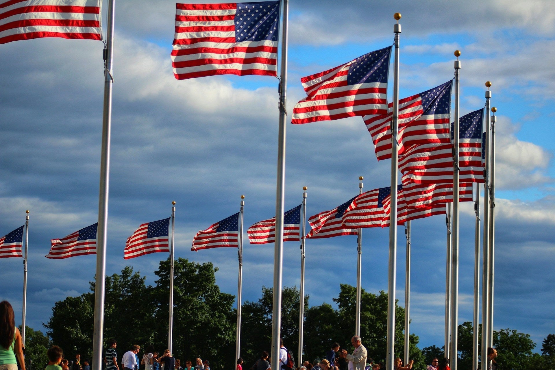 multiple USA flags on tall flagpoles waving in the wind with people gathered below